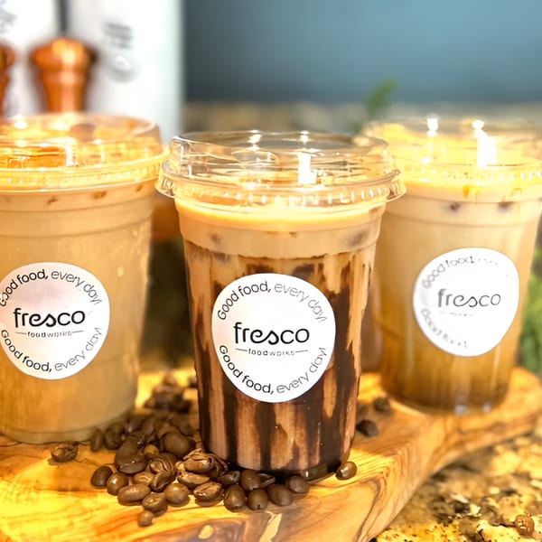 Three iced coffee drinks from Fresco Foodworks, one with chocolate syrup, on a wooden board with coffee beans.