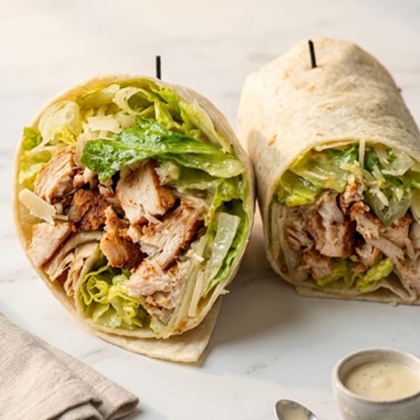 Two chicken Caesar wraps, one cut open, filled with chicken, lettuce, and parmesan cheese.