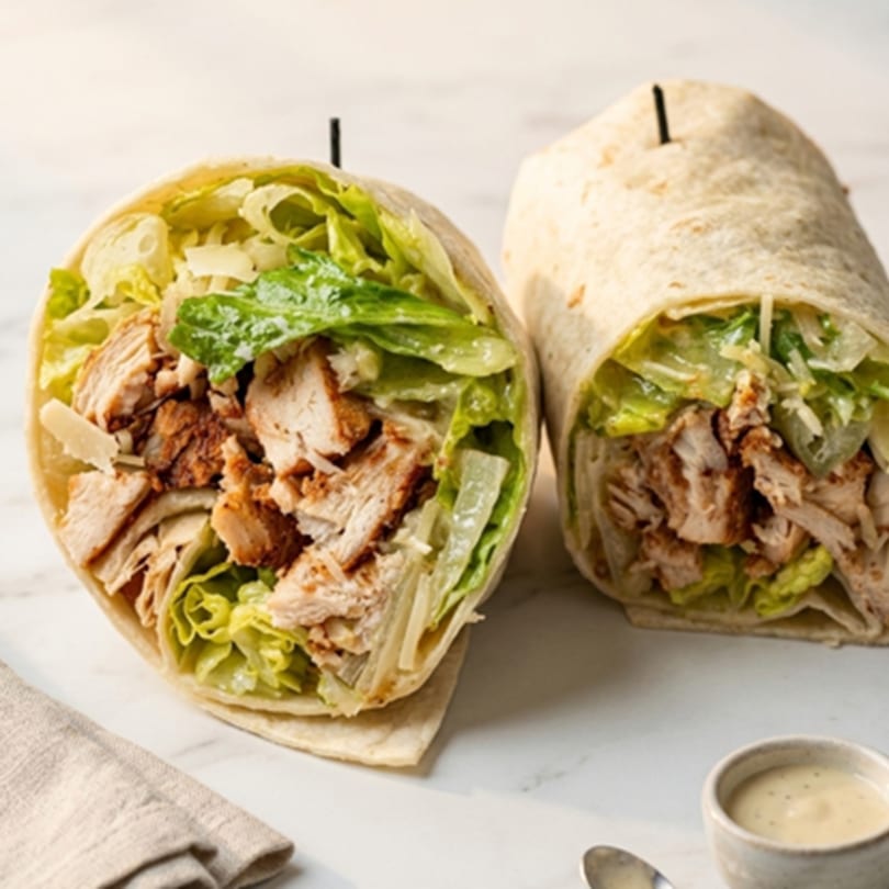 Two chicken Caesar wraps, one cut open, filled with chicken, lettuce, and parmesan cheese.